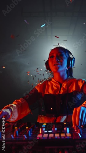 Slow motion vertical video of Asian female DJ in yellow jacket celebrating music moment, dancing and spinning turntable with golden confetti flying in smoky night club atmosphere
