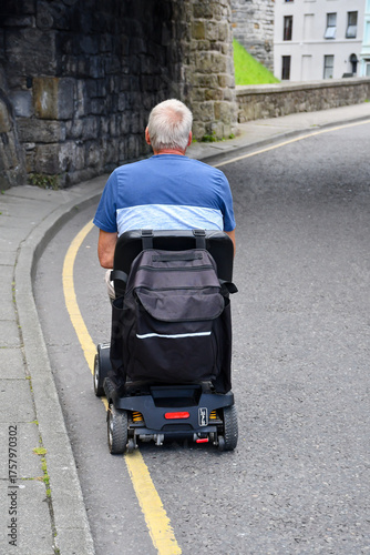 A senior male rides his mobility scooter along the road way, risking danger as the pavement is too narrow for him to drive the vehicle along.