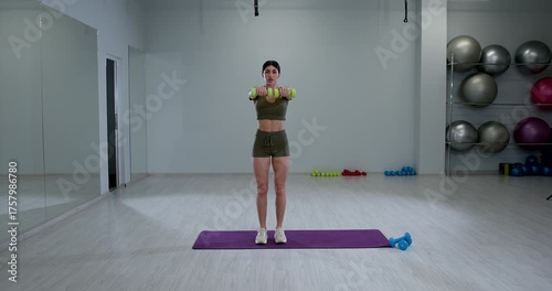 athletic woman stands on a mat in a gym lifting dumbbells in front of her to perform front raises. She focuses on her strength training in a bright spacious environment.