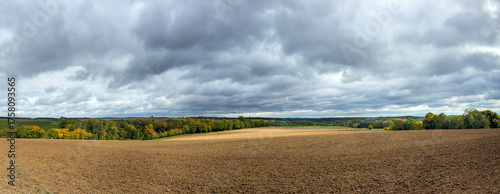 Panoramic view of rural landscape with field and autumn colored forest under cloudy sky