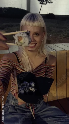 Blonde woman enjoying takeaway sushi with chopsticks at night
