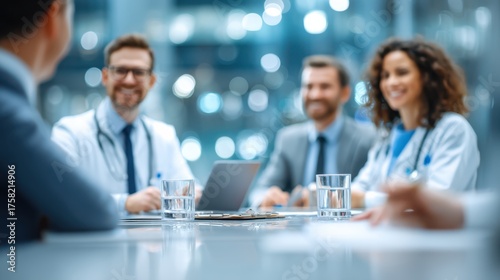 A group of medical professionals in a meeting, discussing work around a table with laptops and glasses of water in a bright, modern office setting.