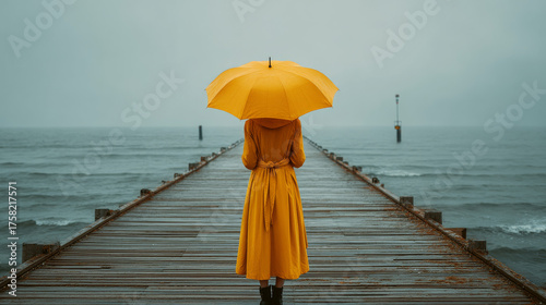 A woman in a yellow raincoat holding a matching umbrella stands on a seaside pier during a storm.