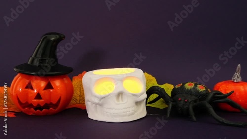 A Halloween composition featuring a skull and a jack-o'-lantern on a dark purple background. A hand holds a candle