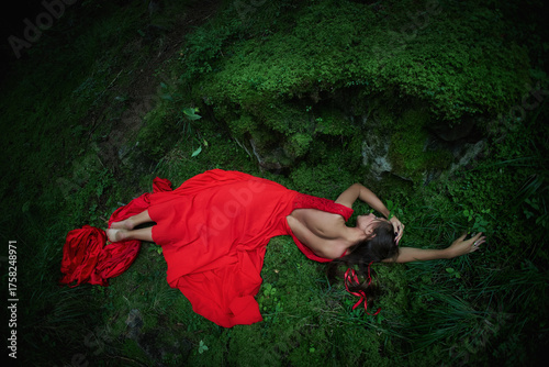 woman lies gracefully on lush green moss in a dimly lit forest. She wears a flowing red gown and appears contemplative against the natural backdrop of trees and foliage