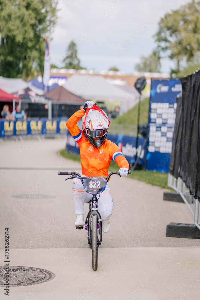 Fototapeta premium Valmiera, Latvia - July 12, 2025: Young boy celebrating victory on BMX bike at cycling event with colorful tents nearby. BMX European championships