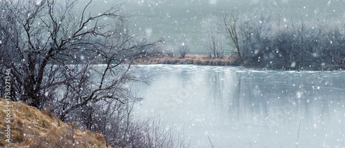 Horizontal rural landscape of the cold season with a frozen river or lake surrounded by bare trees and snowy banks, during a heavy snowfall, creating a gloomy background