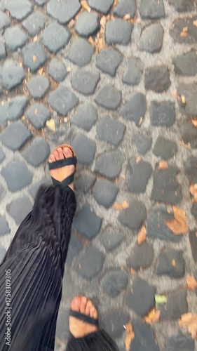 POV shot of female feet with sandals walking on sidewalk or road with cobble stone and leaves on ground. Woman tourist enjoys day outdoors in European city. Travel touring or going to work. Top view