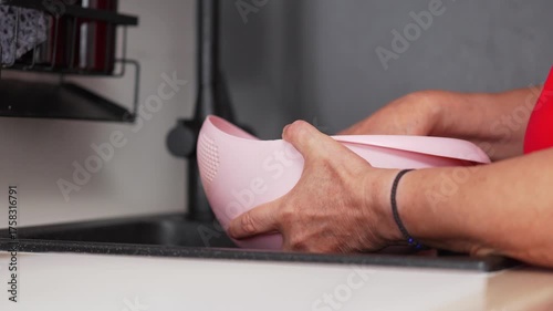 A woman in a red shirt is washing fresh vegetables in a pink colander at the kitchen sink
