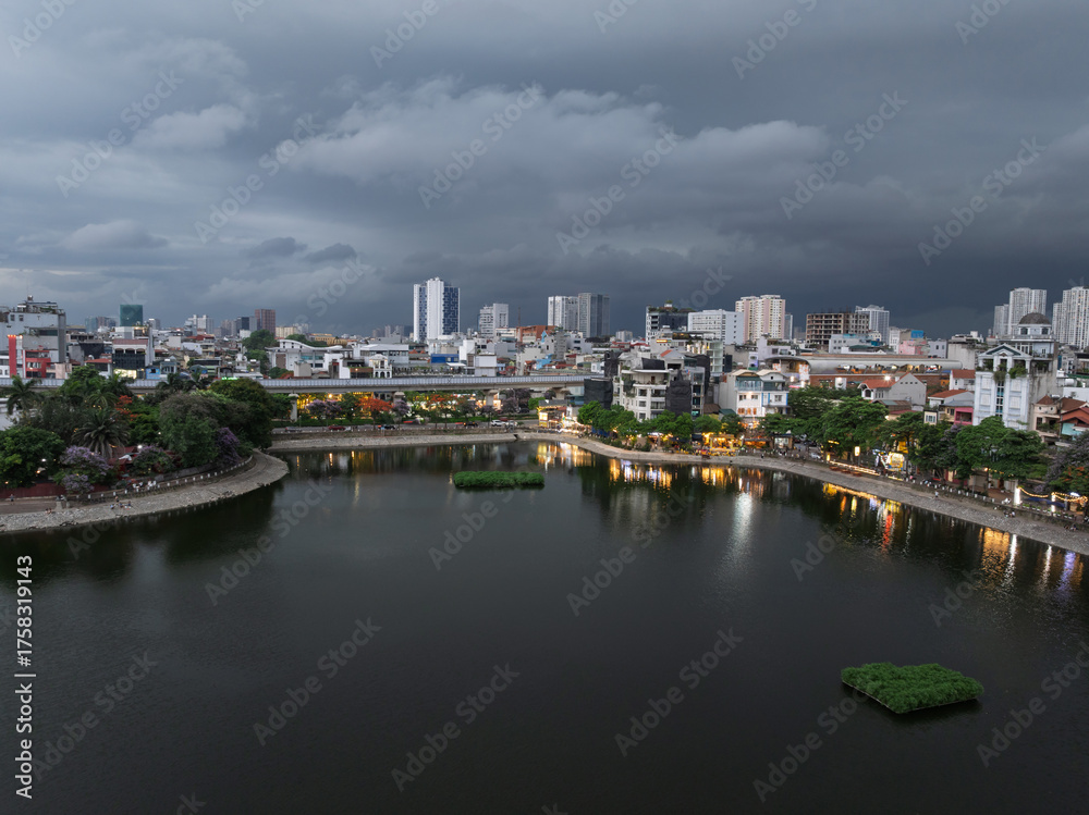 Naklejka premium Evening over Hoang Cau lake with metro line and city lights reflecting under dark clouds.