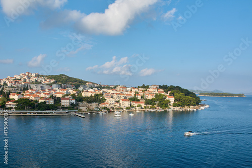 A motorboat leaves a wake on the blue sea as it passes the picturesque coast of Dubrovnik, Croatia, with its red-roofed houses on the hillside.