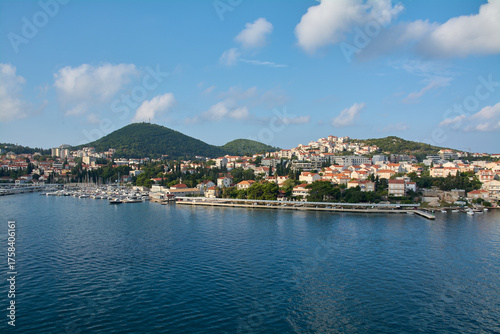 Panoramic view of Gruz harbor in Dubrovnik, Croatia, with boats and yachts in the marina and the city spreading over green hills under a blue sky.