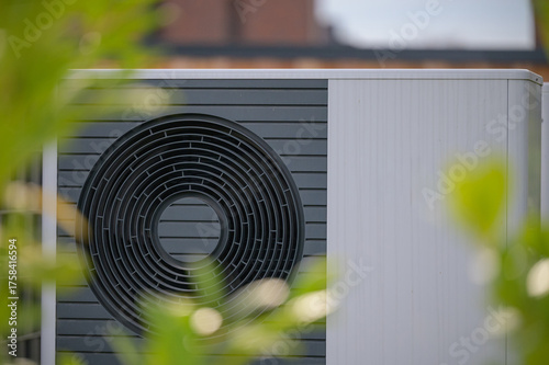 Close-up view of  heat pump unit with a circular fan design framed by soft green foliage. Concept  engineering of sustainable home heating technology. 