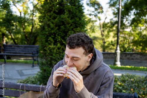 Young man in a brown hoodie enjoys a big bite of his sandwich while sitting on a wooden bench in a sunlit park, surrounded by trees and greenery, with a paper bag beside him