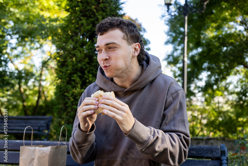 Young man in a brown hoodie sits on a wooden bench chewing a sandwich, smiling with full cheeks, surrounded by greenery and soft sunlight on a relaxed autumn day