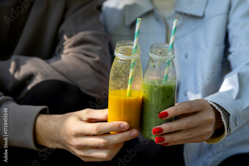 Hands of a man and woman clink together two eco-friendly glass bottles filled with fresh smoothies one vibrant orange and the other green with kiwi seeds each with a striped paper straw, against a sof