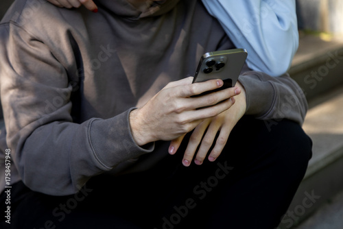 Detail shot of a mans hands holding a modern smartphone as he sits on stone steps, with a womans arm affectionately resting on his shoulder, suggesting a shared digital experience