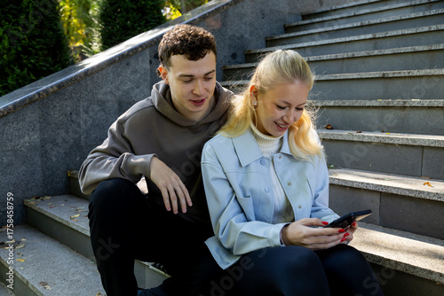 A young man in a brown hoodie and a woman in a light blue jacket share a happy moment while sitting on stone stairs outdoors, smiling as they view something amusing on her phone