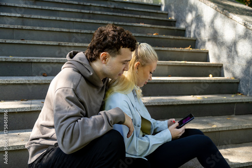 A young woman in a light blue jacket holds a smartphone and looks at it intently, while a young man in a brown hoodie leans in closely to see the screen, both sitting side by side on a staircase in a 