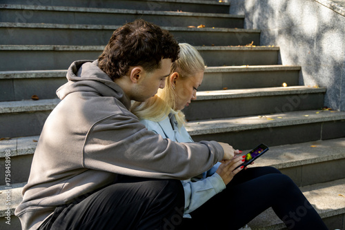 A young man in a brown hoodie points at the screen of a smartphone held by a young woman in a light blue jacket. Both appear focused as they sit closely together on a sunlit stone staircase surrounded