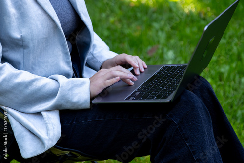 A person in a light blue blazer and dark jeans types on a sleek black laptop while sitting on a chair in a grassy outdoor setting, suggesting remote work or study in a natural environment.