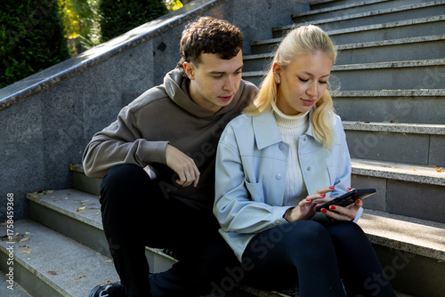 Young man leans in to see the screen as a blonde woman browses her smartphone while sitting on stone steps in a sunny park, sharing a quiet digital moment