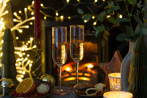 Christmas lights and champagne glasses. Glasses of sparkling wine in warm tone festive atmosphere with blured lights and wood burning stove