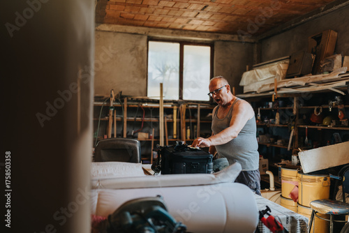 Quadro su tela A middle-aged man in a gray sleeveless shirt and glasses stands at a workbench, organizing tools inside a black bag in a busy, cluttered workshop surrounded by shelves and equipment