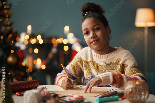 Portrait of Black girl teenager sitting at table writing holiday card with pencil, surrounded by Christmas decorations and craft supplies, Christmas tree in background