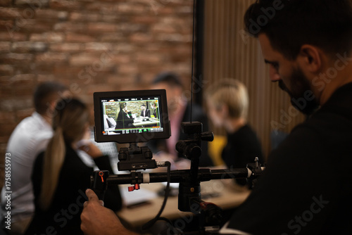 Professional video production setup showing a camera operator monitoring a business meeting being filmed, highlighting teamwork and technology in a modern office.