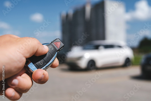 Close-up of a man unlocking a car using a key fob, with a white car parked in a car park in the background.