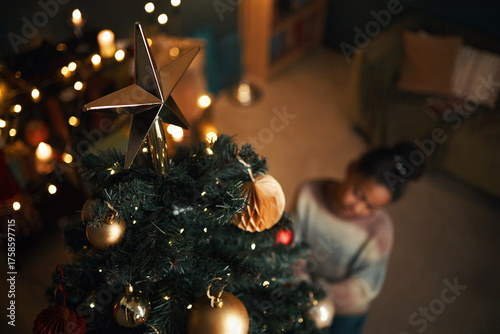 Black child decorating Christmas tree with gold ornaments and lights, standing in warmly lit living room, festive decorations and glowing string lights visible in background