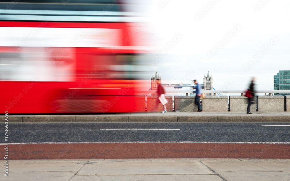 Obraz premium London Rush Hour: A fast-moving red London bus speeds past pedestrians on a pavement near the Thames, with Tower Bridge visible in the background. The motion blur captures the hectic pace of the city.