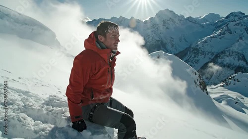 A person in a red jacket, perched on a snow covered mountain, looking down. Sun in the sky