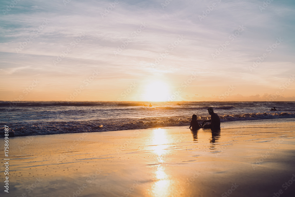 Fototapeta premium Silhouette of a man and child sitting in shallow water at sunset. Warm golden light reflects on the waves, symbolizing family bonding, mindfulness, and the peaceful connection with nature