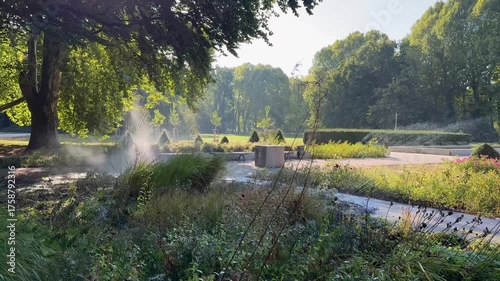 A garden pathway being watered with a hose, sunlight filtering through trees and mist creating a soft rainbow effect in a peaceful park