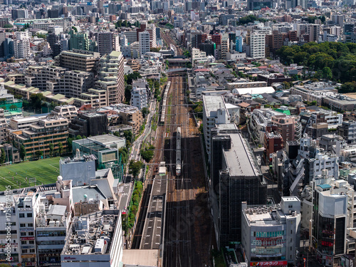 Photography Aerial view shows a Tokyo district as a bullet train passes mid rise blocks, with railway centered, green sports field left, terraced homes, and a wooded park right