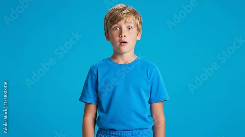 A young, fair-haired boy, surprised with his mouth open, wears a blue shirt against a blue backdrop