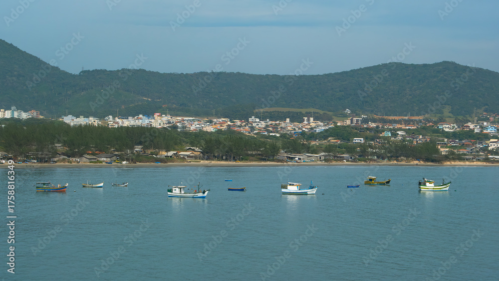 Obraz premium Coastal town scene with colorful fishing boats dotting calm blue waters. A hillside backdrop provides a scenic view of this peaceful seaside community