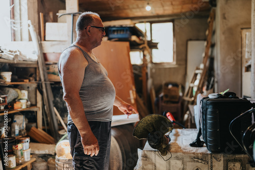 Foto A middle-aged man in a gray tank top and glasses stands in a busy workshop filled with lumber, cabinets, and tools