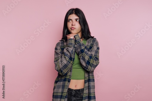 Young woman poses playfully against a pink background wearing a plaid shirt and green top