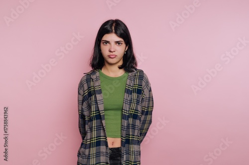 Young woman stands with serious expression in front of pink background wearing casual outfit during indoor photoshoot