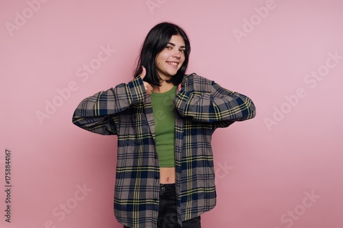 Smiling young woman giving thumbs up in cozy plaid shirt against soft pink background