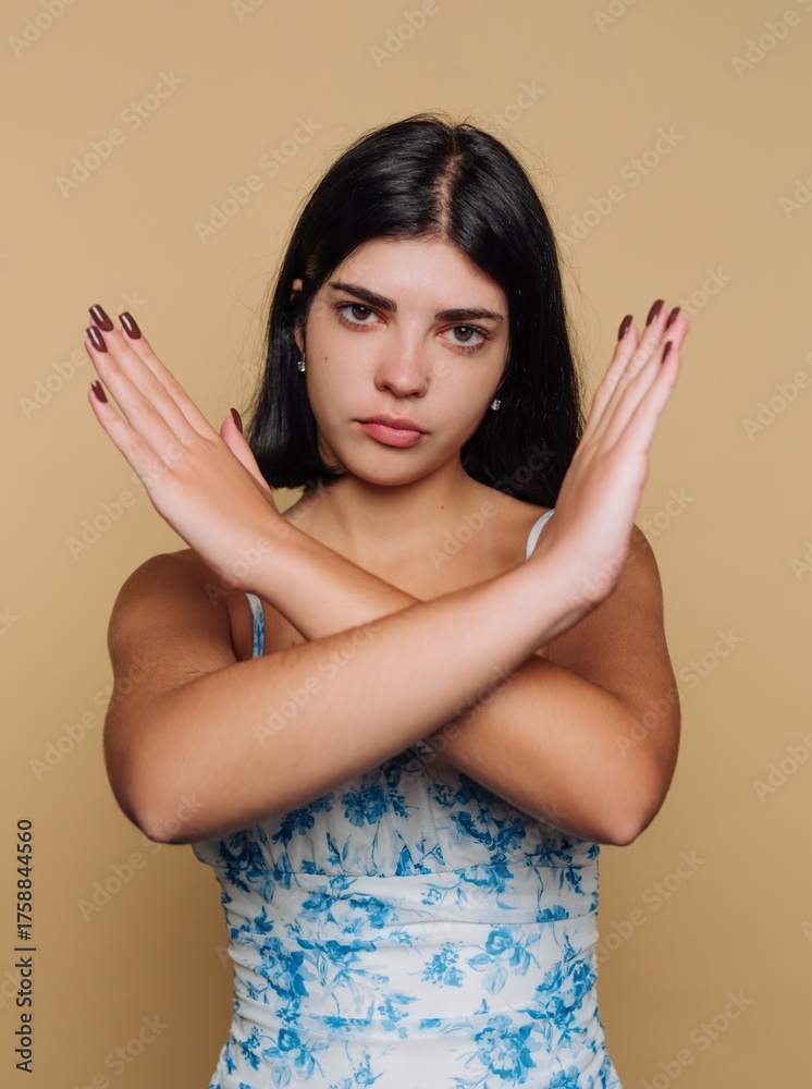Fototapeta premium Young woman in floral dress expresses strong emotions with crossed arms against a neutral backdrop