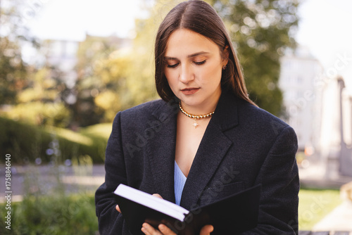 Young student woman freelancer reading book preparing for educational exam at college university outdoors. Female learning studying remotely in city urban area. Woman read book diary outdoor.