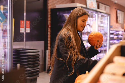 Business smiling long hair woman examining a pumpkin in the produce section of a supermarket. Concept of selecting fresh vegetables, making healthy food choices, and mindful grocery shopping. 