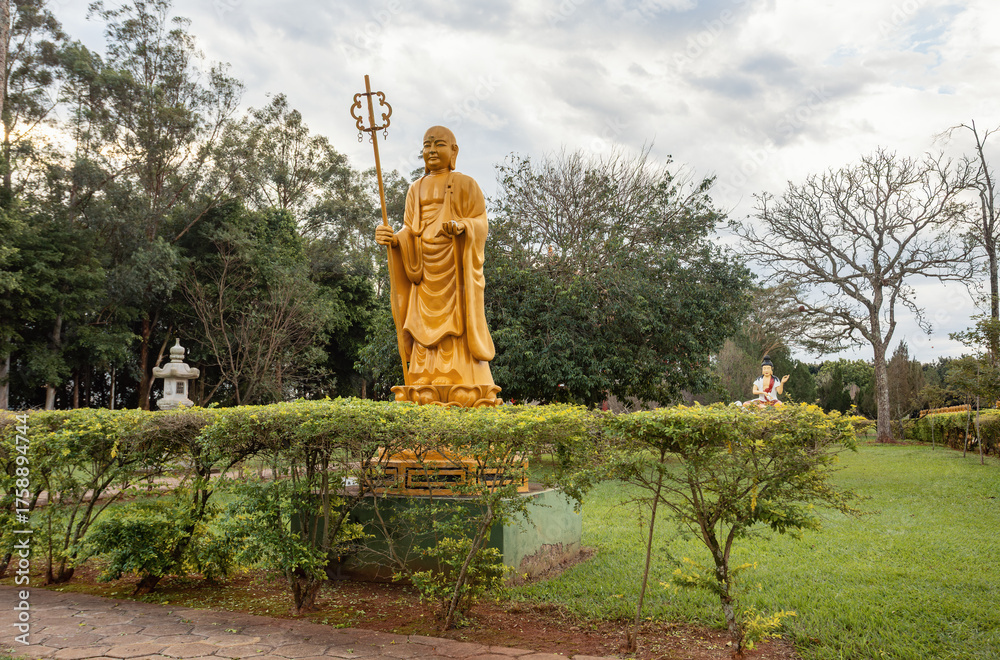 Fototapeta premium The tall, golden statue of Bodhisattva Ksitigarbha, known as the protector of those in hell, stands peacefully in the gardens of the Chen Tien Buddhist Temple in Foz do Iguacu. Brazil.