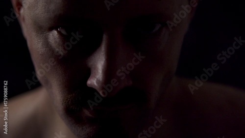 Close-up portrait of an athletic man against a dark background. He looks at the camera with a very intense gaze and moves his facial muscles a little.