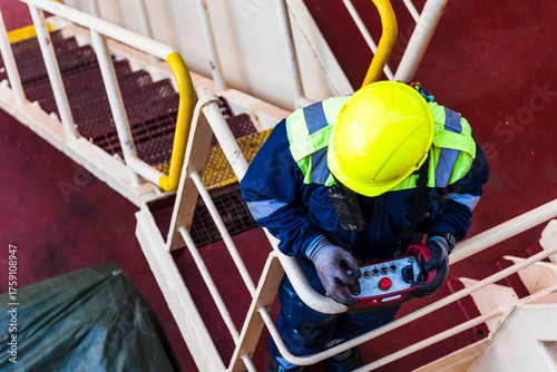 Canvas Print Wearing a yellow hard hat and high-visibility jacket, a seafarer operates a remote control for a provision crane on a merchant ship's deck