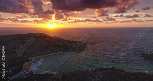 Malta: Golden colorful sunset light painting sky and clouds with vibrant colors over the tranquil ocean water of Ghajn Tuffieha Bay in Malta, breathtaking coastal panorama landscape. Aerial shot